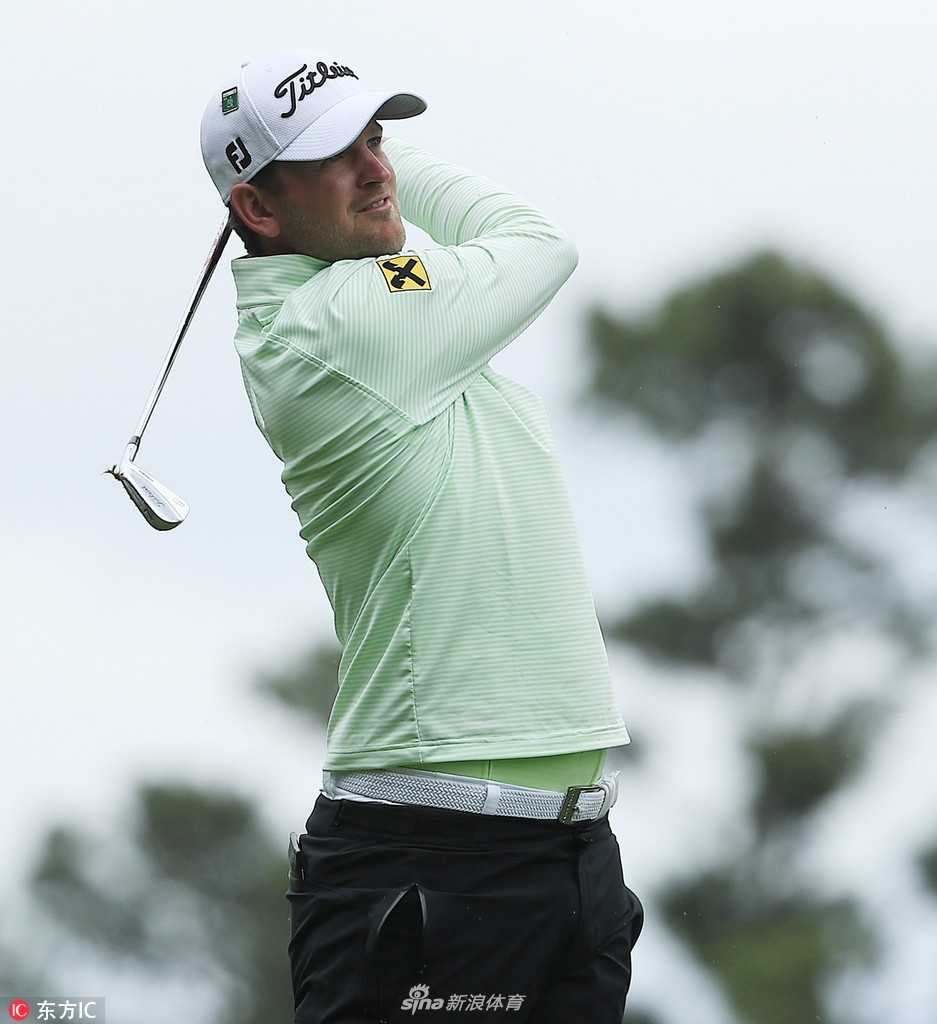 查理·霍夫曼完成挥杆后保持收杆姿势，背景中朦胧的树林如绿色帷幕