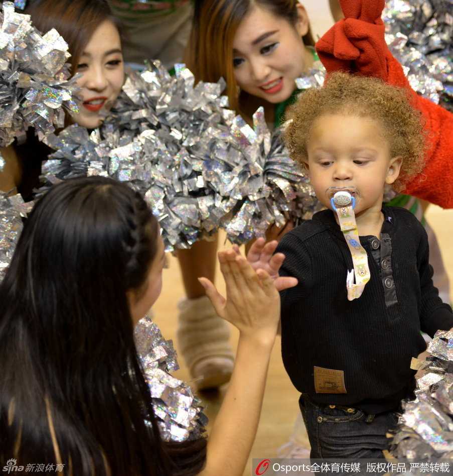 小家伙穿着毛茸茸的连体睡衣，被父亲宽厚的手掌轻轻扶着，微张的小嘴和专注啃手指的模样，简直是把‘萌’字诠释到了极致。