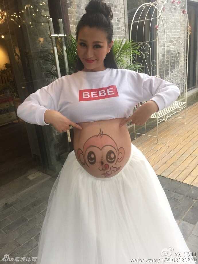 孕妈低头凝视自己的肚皮，上面俏皮的猴子图案与甜美的微笑相映成趣，在江南水乡的柔美背景下，散发出母性的光辉与幸福。