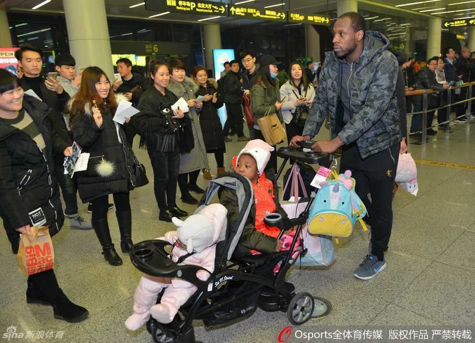 辽篮将士载誉返沈 哈德森小公主萌翻接机现场 哈德森弯着腰,双手稳稳地推着婴儿车,妻子穿着白色羽绒服跟在身侧,一家三口在人群中缓慢穿行,画面温馨有爱。