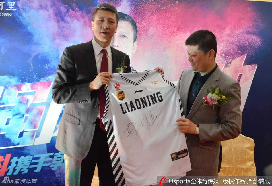 郭士强手里拿着一件辽宁男篮的绿色球迷服，正低头为球迷签名，他神情专注而认真，旁边的球迷们举着手机记录这一时刻