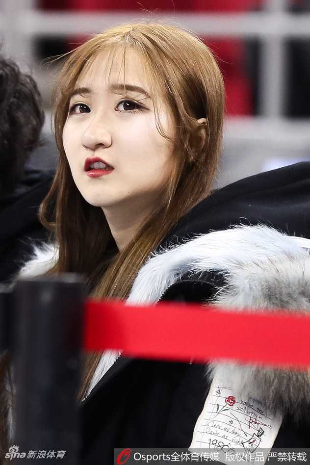 一位身着浅灰绒衣的温婉女球迷坐在看台上，眼神专注而柔和，仿佛整个球场的喧嚣都与她无关，她的眼里只有那一道橘红色的弧线。