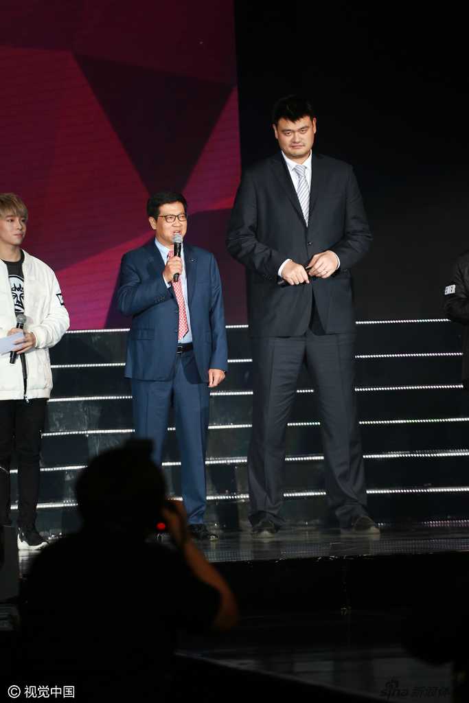 姚明与现场的音乐人及嘉宾并肩而立，巨大的身高差构成一幅有趣的画面，他正侧耳倾听，眼神中流露出对音乐世界的尊重与好奇。