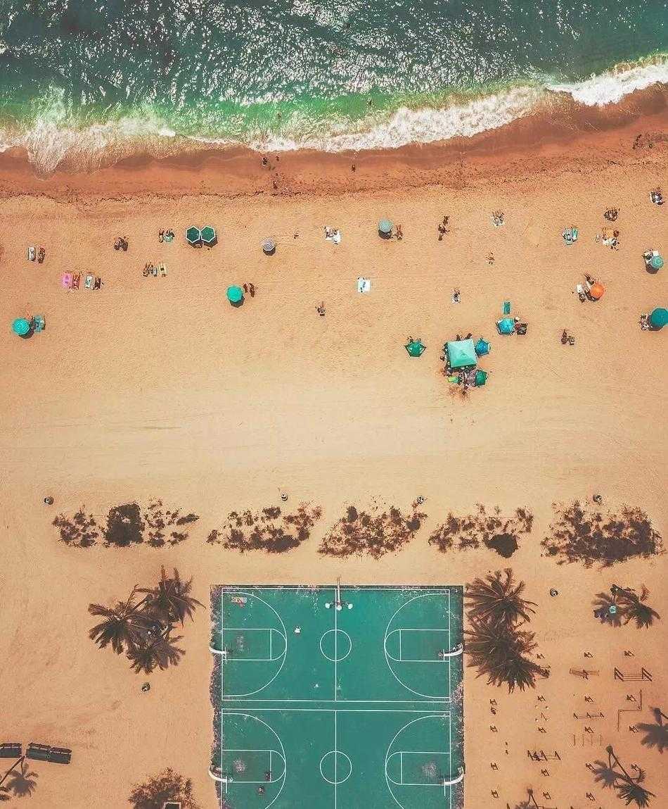 俯瞰光影交错,探寻篮球场的几何诗意 黄昏时分,几名少年在露天篮球场上打球,暖黄的灯光将他们包围,场边的绿树和远处的高楼剪影构成了宁静而充满活力的生活画卷。