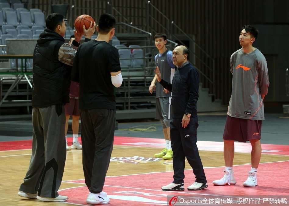 八一队年轻内线球员在篮下进行对抗训练，表情坚毅，肌肉线条在汗水浸润下闪闪发光，王治郅在一旁双手叉腰，目光如炬地注视着每一次起跳和出手，准备随时上前指导。