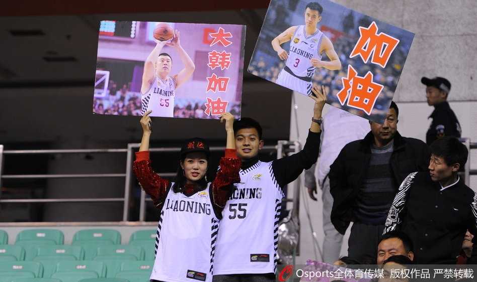 看台上一对父子球迷，父亲满脸笑容地抱着年幼的儿子，儿子手中也拿着助威棒。身后无数双手臂高举，人声鼎沸，父子俩共同享受着篮球带来的快乐时光，这正是球迷文化代代相传的最好写照。