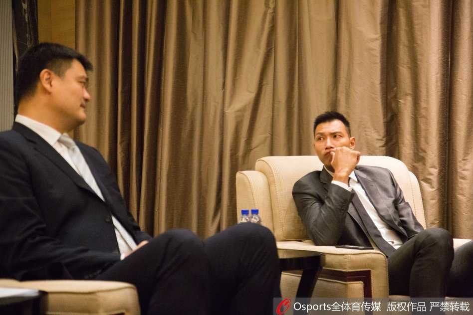 易建联身着休闲西装端坐于主席台前，神情专注而坚定，仿佛已为新赛季的征战积蓄了全部能量。