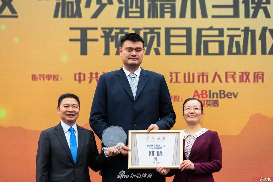 身穿深色上衣的姚明在校园教学楼前，被一群穿着红色校服的学生簇拥着，他面带微笑，亲切地拍着一位男生的肩膀，如同一位温和的邻家大哥哥。