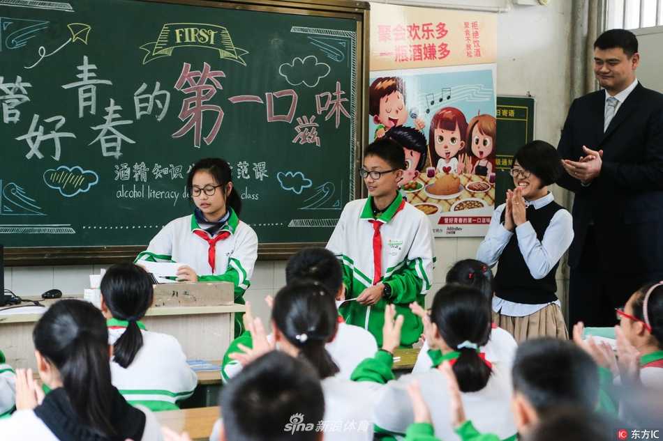 在一个摆满奖杯和装饰的室内空间，姚明弯腰侧身，与一群孩子聚在一起，他的姿态放得很低，几乎与孩子们平视，大家仿佛正在分享一个共同的秘密，画面温暖而平等。