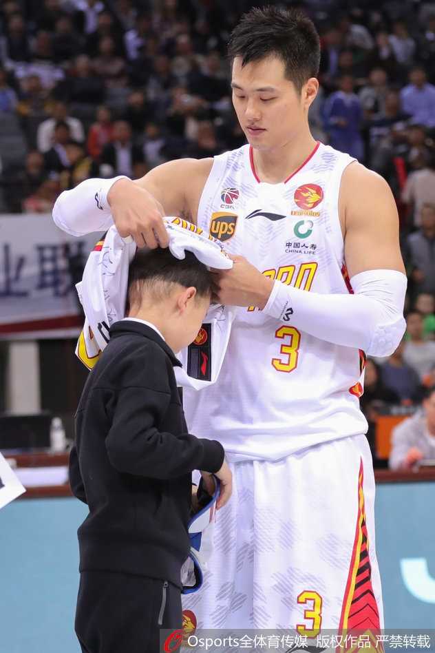 孟铎身穿白色深圳队球衣，弯下腰双手将签名球衣递到轩轩面前，他的脸上带着亲切的笑容，轩轩则伸出双手准备接过这份珍贵的礼物，两人眼神交汇的瞬间充满温情。