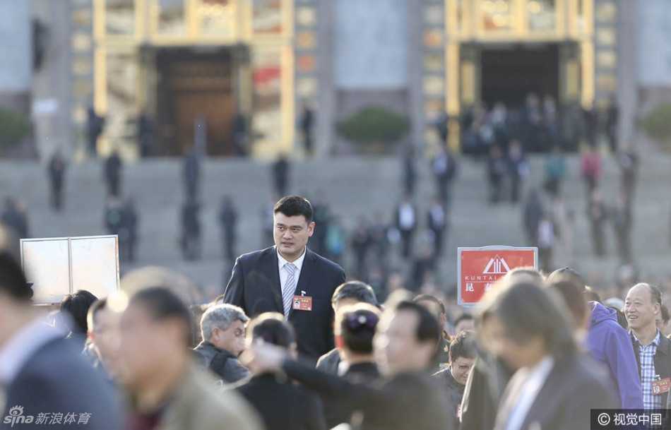 姚明独自行走在大会堂外的台阶上，背景是庄严的国徽与飘扬的红旗，他以仰望的姿态看向远方，仿佛在审视着体育强国的未来蓝图