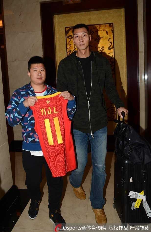 易建联身穿一件简约的灰色T恤，面带微笑，正耐心地为身旁的几位小球迷在篮球和球衣上签名。