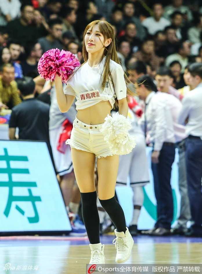 广厦篮球宝贝演绎力与美 赛场热舞点燃萧山战神之夜 马尾辫啦啦队员回眸露出甜美微笑,背景中广厦球员正在热身,刚柔并济的画面定格了赛场内外的和谐共处。