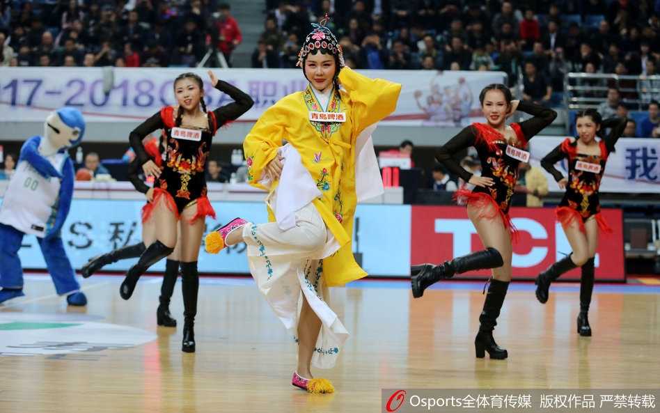 当国粹邂逅篮坛:福建篮球宝贝戏曲服饰演绎别样助威风采 镜头拉近,一位篮球宝贝的妆容精致细腻,额头贴着戏曲特有的水钻,眼神中透露出既属于演员的英气又属于啦啦队员的活力,背景是模糊的球场灯光。