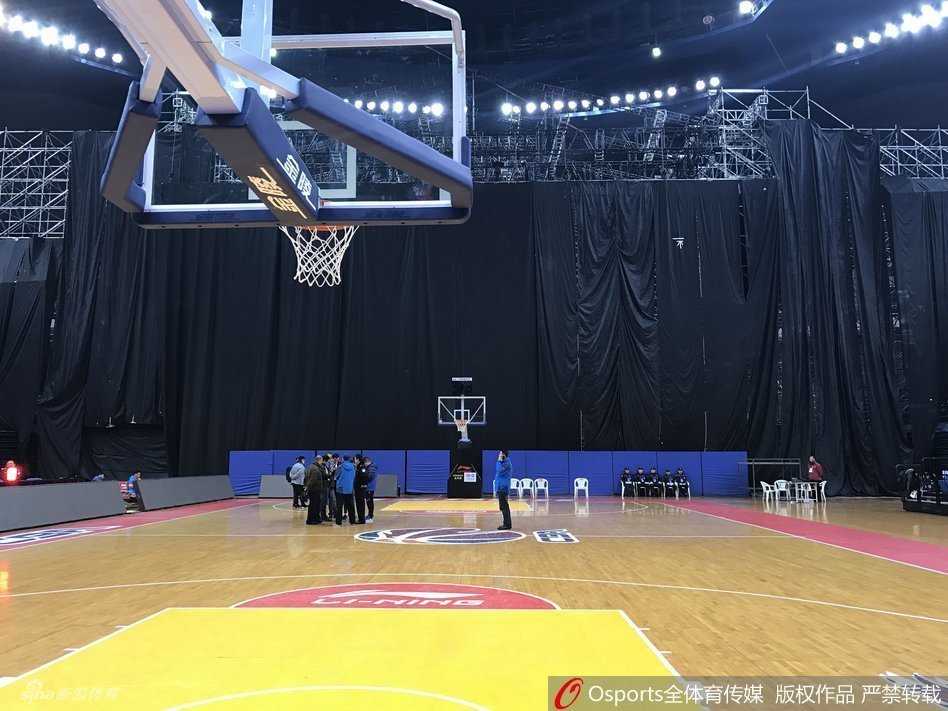 当篮球架遇见演唱会舞台:一场因“跨界”而取消的CBA比赛 几名身穿工作服的工人正在舞台的脚手架旁进行最后的检查,他们身后是被黑布严密遮盖的舞台主体,背景里青岛主场的冠军旗和退役球衣横幅若隐若现,现实与荣誉在此刻形成鲜明对比。