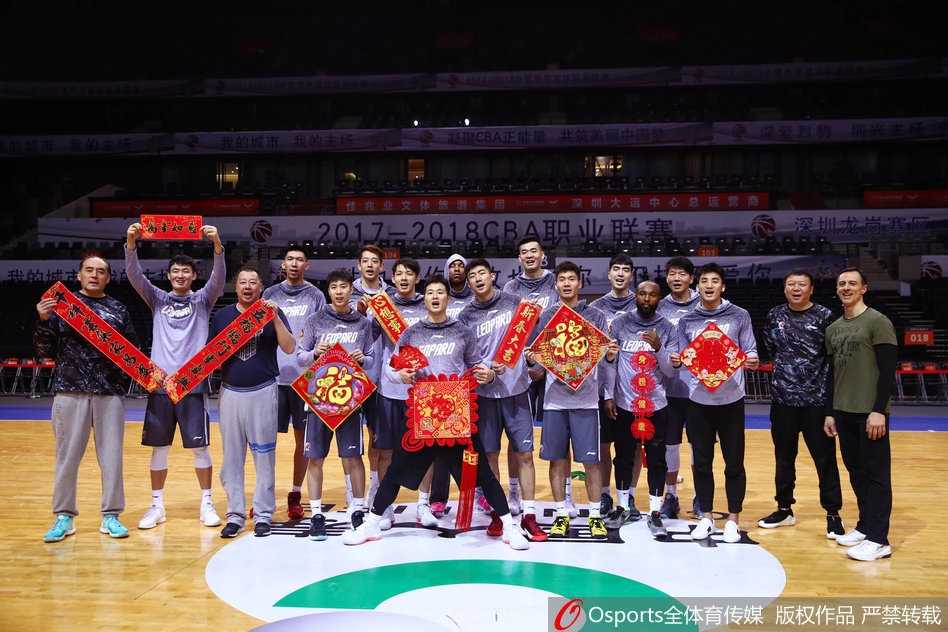 主教练王建军指导在拍摄现场露出难得的轻松微笑，他与外援萨林杰并肩而立，两人手中拿着春联，仿佛在战术板上画出了一幅新春取胜的蓝图。