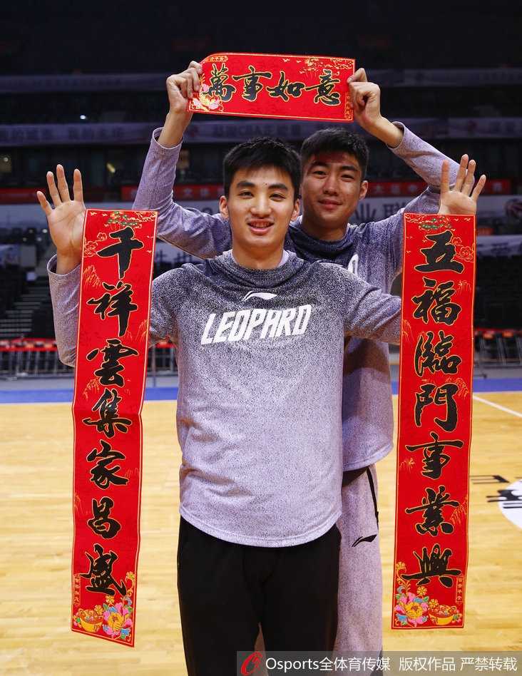 深圳男篮队长孟铎在喜庆的红色背景下，双手紧握鼓槌奋力敲响铜锣，眼神中充满坚定与笑意，仿佛在为即将到来的新春敲开胜利之门。