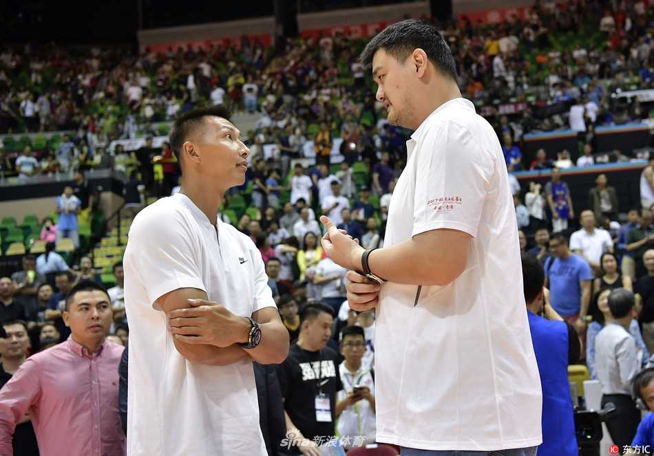 易建联与姚明相邻而坐，共同注视着场上的小朋友球员。阿联一只手轻轻托着下巴，嘴角带着轻松的笑意，姚明则双手交叉放在身前，两位中国篮球的领航者此刻更像两位慈祥的守护者。