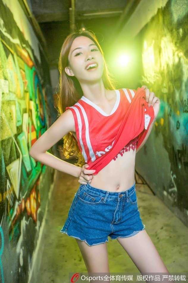 一位身材曼妙的中国女球迷，身穿紧身白色上衣与牛仔裤，在火箭队主场看台前自信地展示着姣好身段，她手举手机仿佛正在记录这一刻为周琦加油的独特心情。