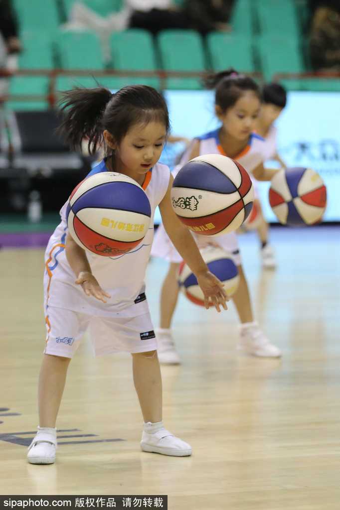 一个小女孩在表演中不慎滑倒，但她迅速爬起来，顾不上拍身上的灰尘，立刻找回节奏继续拍球，脸上依旧挂着甜甜的笑容，萌态可掬又惹人疼爱。