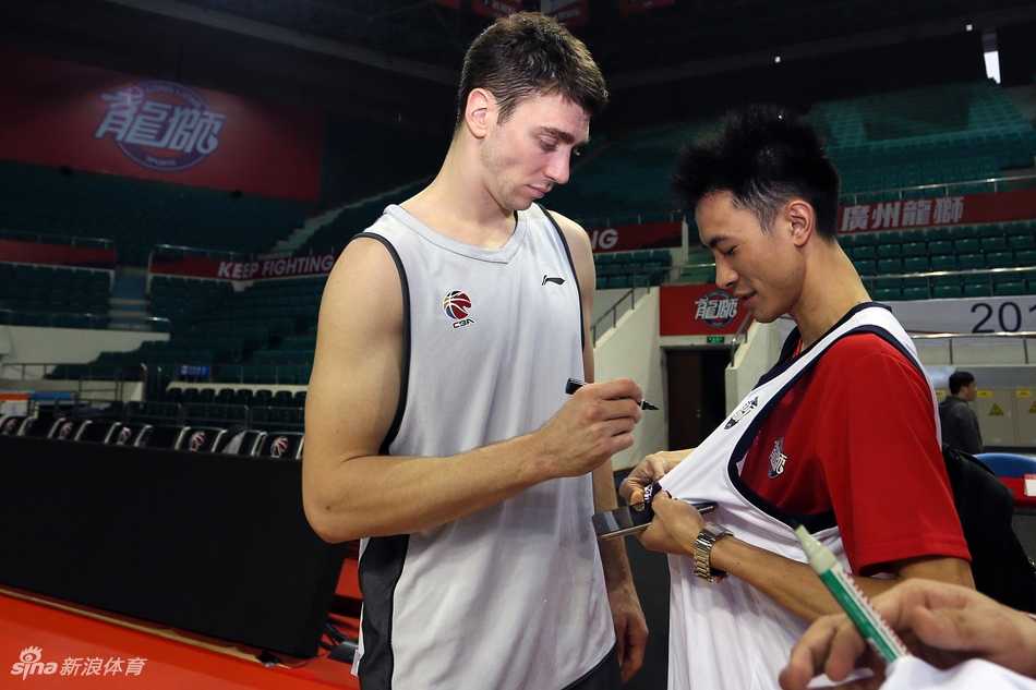 羊城冬日里的热血相聚:广州男篮球迷开放日侧记 广州男篮开放日的趣味对抗环节,一名球员高高跃起试图封盖队友的投篮,旁边其他的队友和场边的球迷都被这一瞬间逗乐,气氛欢快。