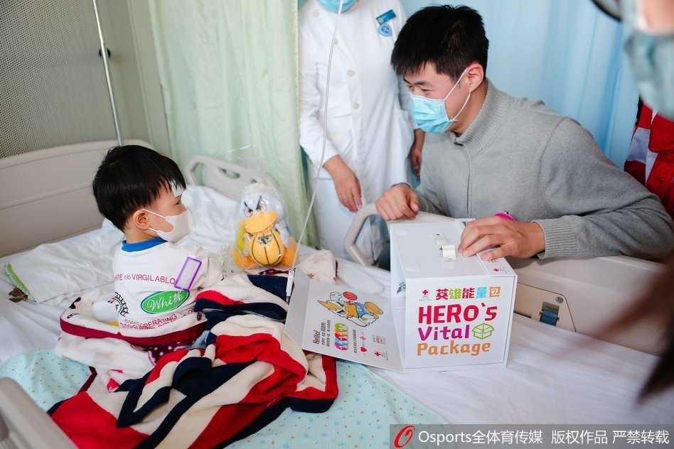翟晓川张松涛与白血病儿童共绘温暖篮球梦 翟晓川、张松涛和吉祥物霹雳鸭一起,手捧着装满礼物的英雄能量包,准备走进病房送给孩子们,他们的脸上洋溢着温暖而真诚的微笑,背景是贴满卡通图案的医院走廊