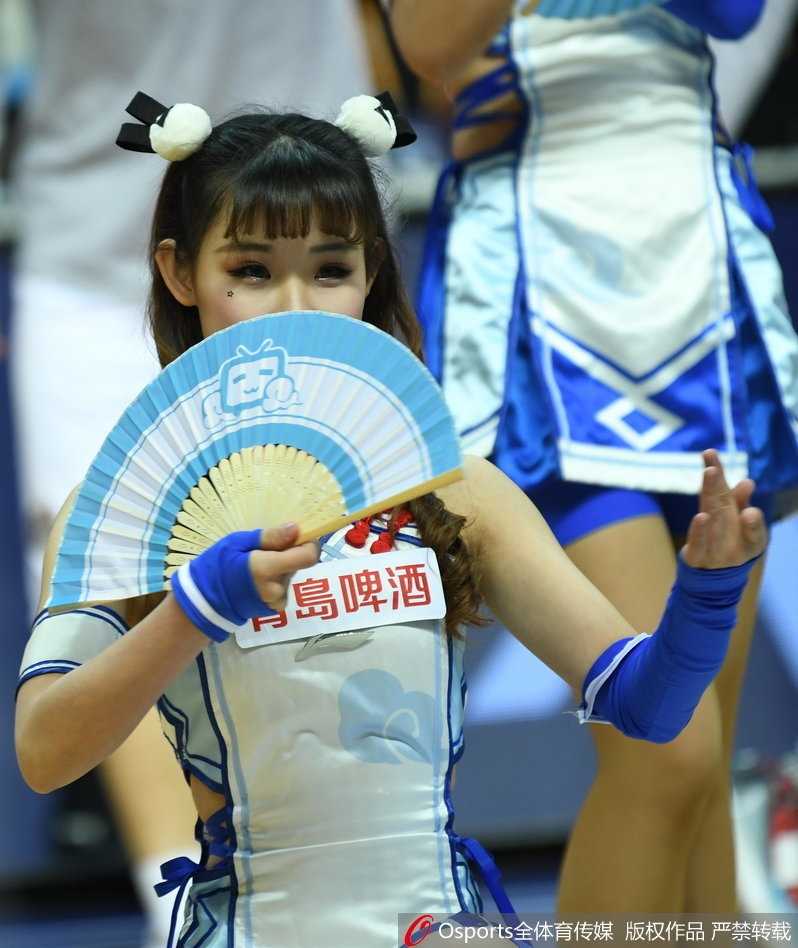 萌酥了!上海滩的二次元旋风与一场属于硬汉的加时悲喜剧 一位啦啦队女孩的特写,她微微侧身,对着镜头露出治愈系的甜笑,手上拿着一个印有可爱卡通形象的应援手幅,背景是虚化的球场和观众,整个画面充满了青春与活力的气息。