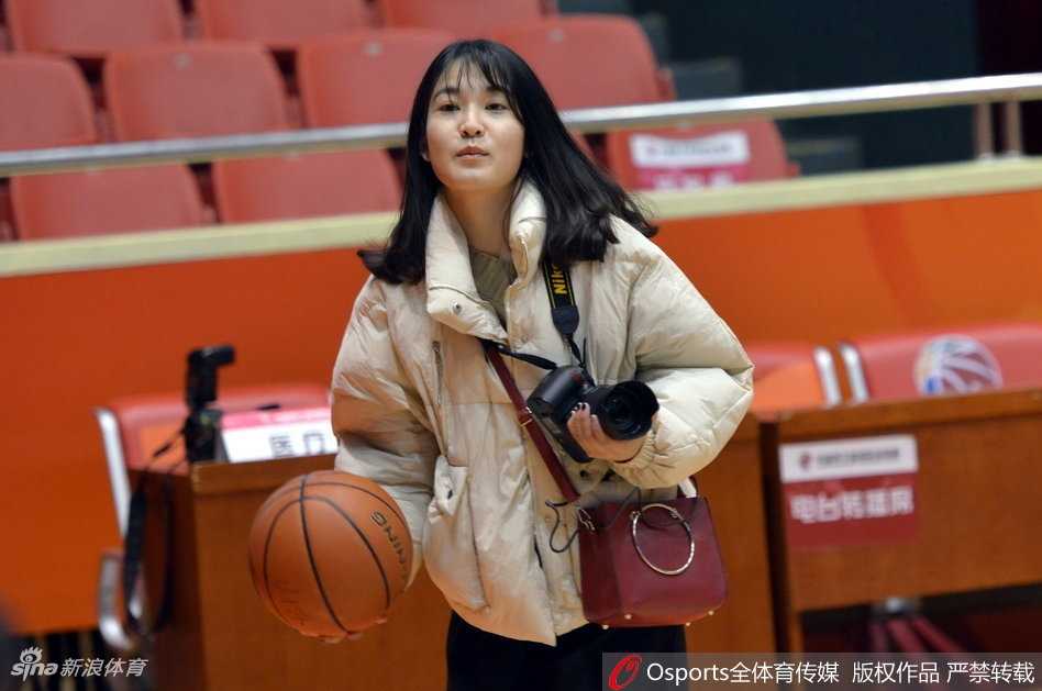 训练场全景，两边的篮架下分别摆放着新疆和山东队的训练用球。美女记者背对镜头，提着采访包正走向出口，整个球馆在灯光照射下显得格外庄重，一天的探营报道圆满结束。