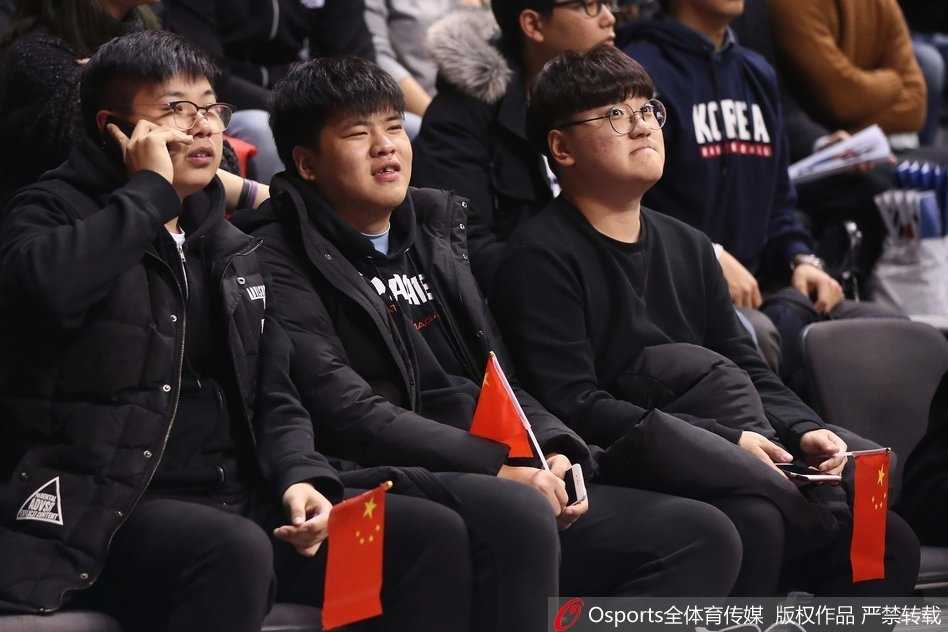 赛场边的看台上，一名年轻的中国球迷脸上洋溢着自豪的笑容，他身体前倾，奋力挥舞着手中的小型五星红旗，旗帜的红色与他脸上的兴奋交相辉映。