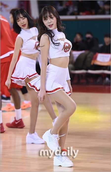 長髮飄逸的啦啦隊員身著白色緊身露臍裝，搭配藍色超短裙，在聚光燈下盡情舞動。她曼妙的身材曲線在動感舞步中展露無遺，彷彿是賽場上跳動的精靈。