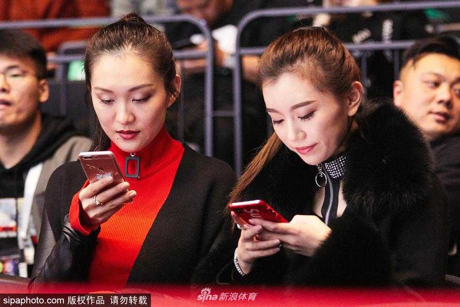 丁彦雨航女友举起手机对着球场拍照，背景里是模糊的观众和璀璨的场馆灯光，她的侧脸轮廓柔和，正试图透过小小的取景框，将男友在赛场上的高光时刻定格为永恒的记忆。